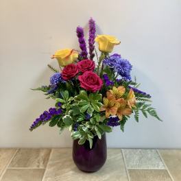 Mixed bouquet of roses, purple blooms, and orange flowers in a purple vase