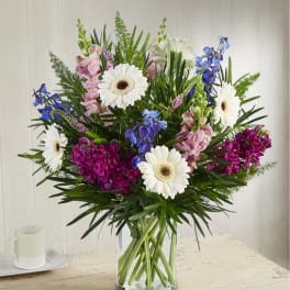 Mixed bouquet of white gerbera daisies, pink blooms, and blue flowers in a glass vase
