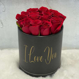 Black hatbox of red roses with gold 'I Love You' text, displayed on white fur.