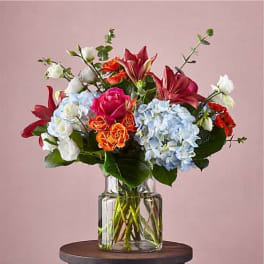 Mixed bouquet of blue hydrangeas, red lilies, and roses in a clear glass vase on a small round table.