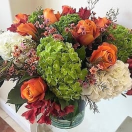 Low glass vase of orange roses, green and white hydrangeas, and red accent flowers.