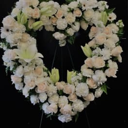 Heart-shaped white floral wreath with pale roses and lilies on a black stand