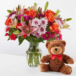 Mixed bouquet in a glass vase beside a teddy bear with a red bow