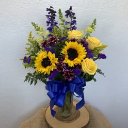 Bouquet of yellow roses, sunflowers, and purple flowers in a glass vase with a blue ribbon