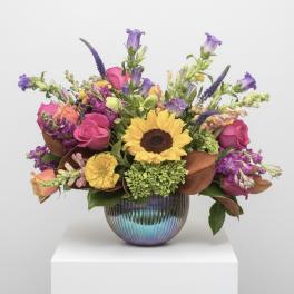 Mixed bouquet with a sunflower, pink roses, and purple blooms in a ribbed iridescent vase