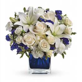 Compact white lilies, roses, and carnations with blue accents in a blue glass cube vase