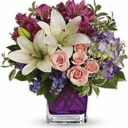 Mixed bouquet of white lilies, pink roses, and purple blooms in a purple glass cube vase