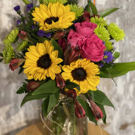 Bouquet of yellow sunflowers, pink roses, and purple filler flowers in a glass vase