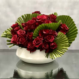 Low arrangement of dark red roses with fan-shaped green foliage in a white ceramic bowl