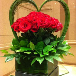Red roses arranged in a heart-shaped display with green foliage in a square vase