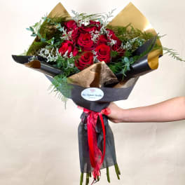 Hand-tied bouquet of red roses with white filler flowers wrapped in black and gold paper and red ribbon