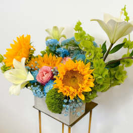 Bright mixed bouquet with sunflowers, lilies, roses, and blue filler flowers in a square vase