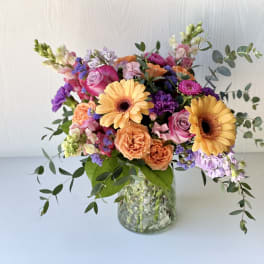 Mixed bouquet of orange gerbera daisies, pink roses, and purple flowers in a glass vase