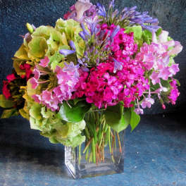 Mixed bouquet of pink blooms, green hydrangeas, and purple flowers in a clear glass cube vase