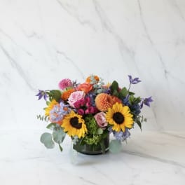 Mixed bouquet with sunflowers, roses, and purple blooms in a low vase