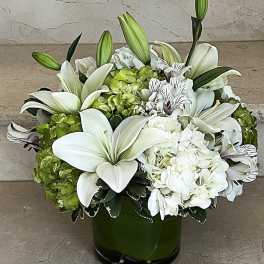 Low arrangement of white lilies, hydrangeas, and alstroemeria in a green glass cylinder vase