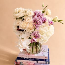White roses, hydrangeas, and orchids arranged in a glass vase on stacked books