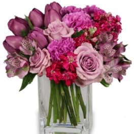 Compact arrangement of pink and purple roses, tulips, and mixed blooms in a clear glass vase