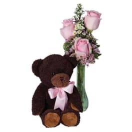 Brown teddy bear with pink bow beside a tall green vase of light pink roses and small white blossoms