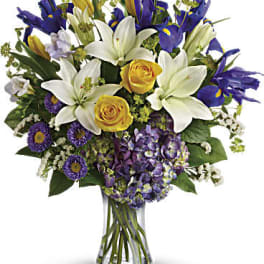 Tall vase arrangement with white lilies, yellow roses, purple irises, asters, and hydrangeas