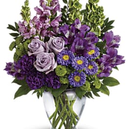 Tall arrangement of purple flowers with lavender roses and green bells of Ireland in a clear glass vase