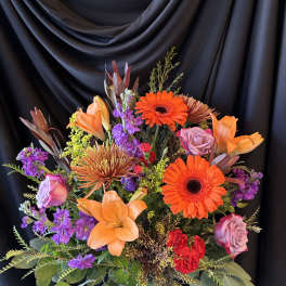 Bright mixed bouquet with orange lilies, gerbera daisies, and purple roses in a glass vase