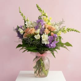 Mixed bouquet of purple, peach, and white flowers in a glass vase with a pink ribbon.