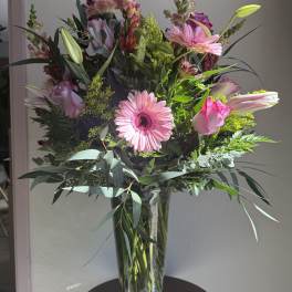 Tall arrangement of pink gerbera daisies, roses, lilies, and snapdragons in a clear glass vase