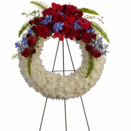 Circular funeral wreath with red roses, white flowers, and blue accents on a stand