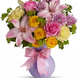 Bouquet of pink lilies, roses, and yellow carnations in a lavender vase with a pink bow