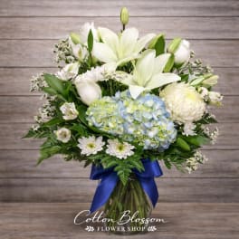 White lilies and blue hydrangeas in a glass vase with a blue ribbon
