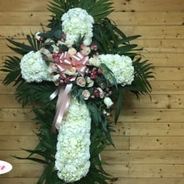 White floral cross arrangement with pink roses and ribbon on a wooden wall