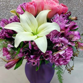 Pink roses and purple flowers arranged in a purple vase with a white lily