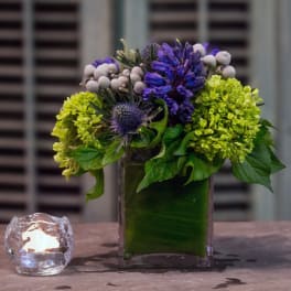 Purple and green floral arrangement in a square glass vase beside a lit candle.