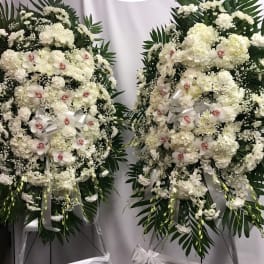 Two large white floral standing sprays with ribbons and dark green foliage