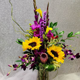 Tall mixed arrangement with sunflowers, purple orchids, yellow calla lilies, and protea in a clear glass vase.