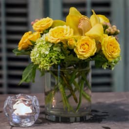 Yellow roses and orchids arranged in a clear glass vase beside a lit candle