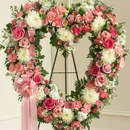 Heart-shaped standing wreath of pink roses, carnations, and white flowers with a pink ribbon bow