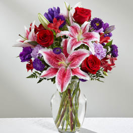 Bouquet of pink lilies, red roses, and purple flowers in a clear glass vase