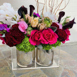Two mirrored vases hold a colorful floral arrangement with roses and calla lilies.