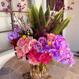 Pink roses and purple orchids arranged in a glass vase with tall green leaves.