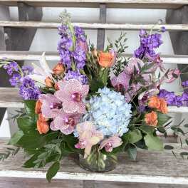 Mixed bouquet with purple blooms, orange roses, blue hydrangea, and orchids in a glass vase