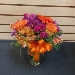 Colorful bouquet of roses, alstroemeria, and purple daisies in a glass vase with an orange ribbon