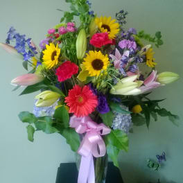 Mixed bouquet with sunflowers, lilies, gerbera daisies, and a pink ribbon in a glass vase