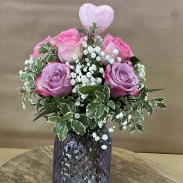 Pink roses with baby's breath in a purple glass vase, topped with a heart pick