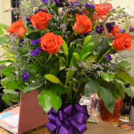 Orange roses in a glass vase with purple filler flowers and a ribbon bow