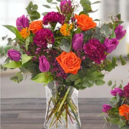 Bouquet of orange roses, purple tulips, and magenta flowers in a clear glass vase