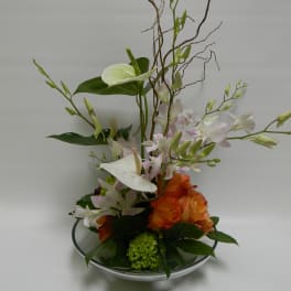 Tall floral arrangement with white orchids and orange roses in a shallow glass bowl