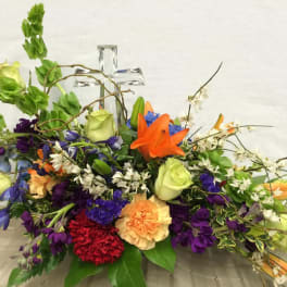 Colorful mixed flower arrangement with roses, lilies, and carnations around a clear glass cross