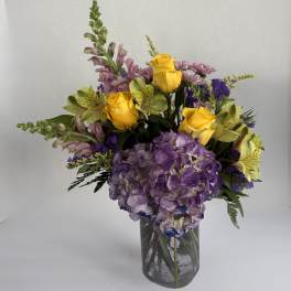 Bouquet of yellow roses, purple hydrangea, and mixed blooms in a glass vase